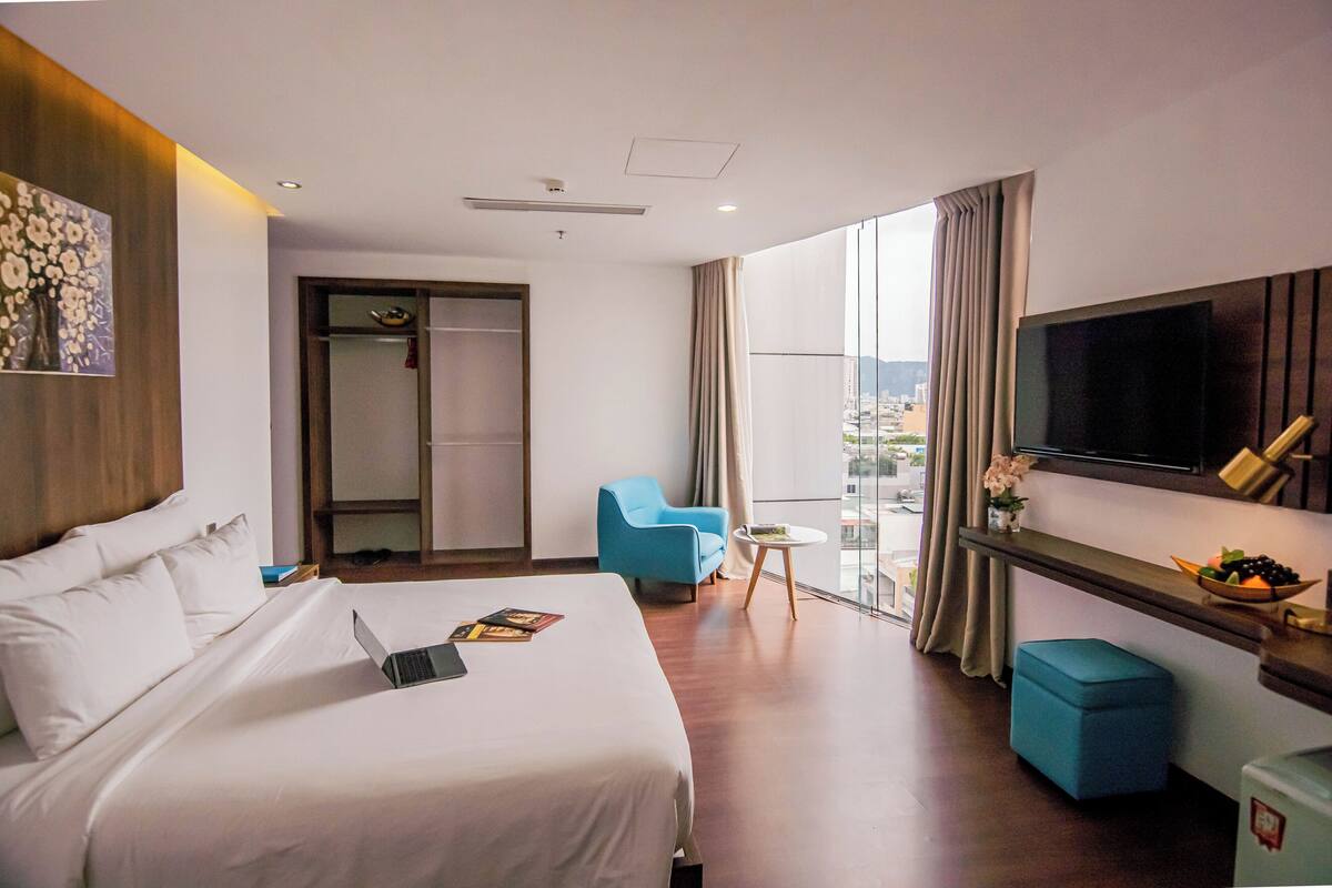 A hotel room with a bed, a desk, a chair, a TV, and a window with a city view.