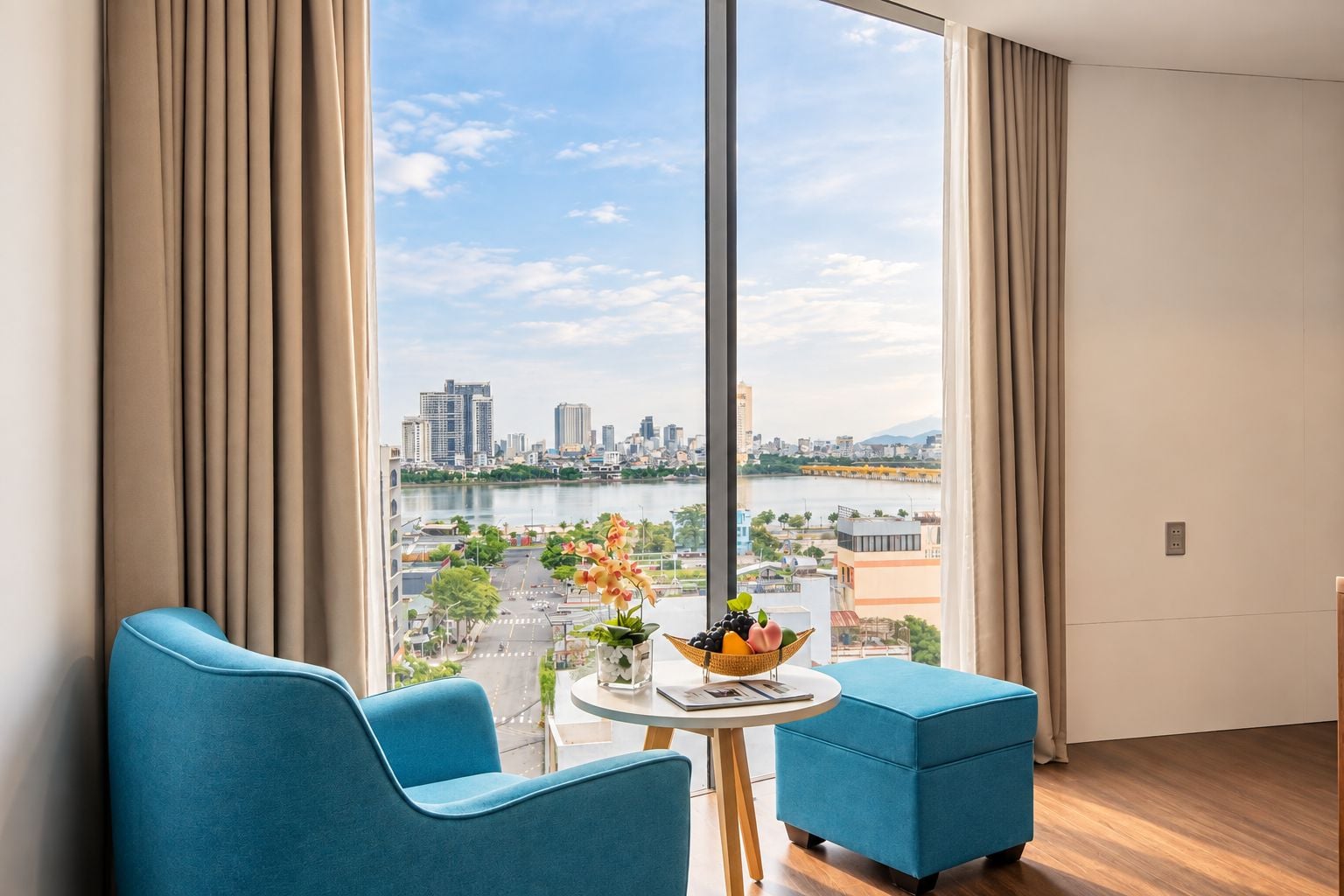 Une chambre moderne dotée d’une grande fenêtre, de deux fauteuils bleus, d’une petite table ronde sur laquelle se trouve un panier de fruits, et qui offre une vue sur le paysage urbain et la rivière.