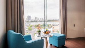 A modern hotel room with a large window offering a city view, a round table with a fruit bowl, and two blue armchairs. - Gold Central Hotel by Haviland (Da Nang)
