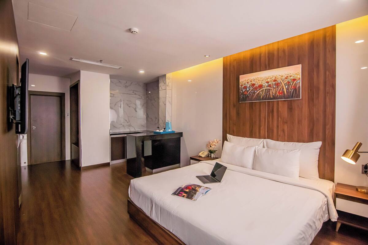 A hotel room with a large bed, a desk with a laptop, a painting on the wall, and a wooden panel design.