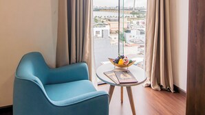 A modern hotel room with a large window, a blue armchair, a small round table with a fruit bowl, and a view of a bridge and cityscape. - Gold Central Hotel by Haviland (Da Nang)