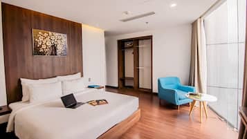 A hotel room with a large bed, a laptop, a blue armchair, a small table, and a large window.