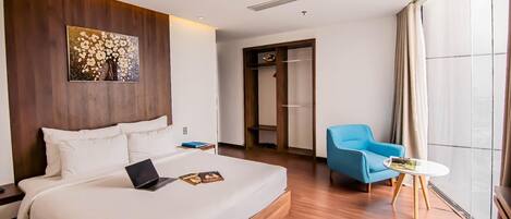 A hotel room with a large bed, a laptop, a blue armchair, a small table, and a large window.
