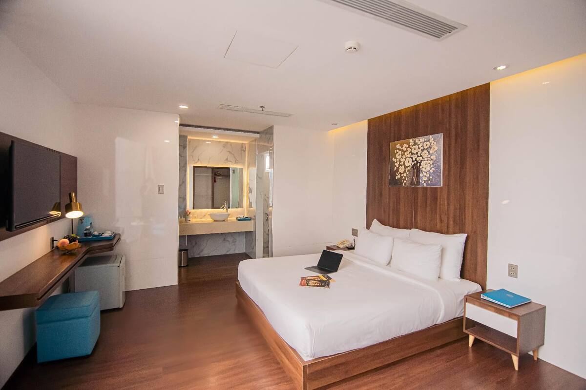 A modern hotel room with a large bed, a desk with a lamp, a small seating area, and a bathroom visible through an open door.