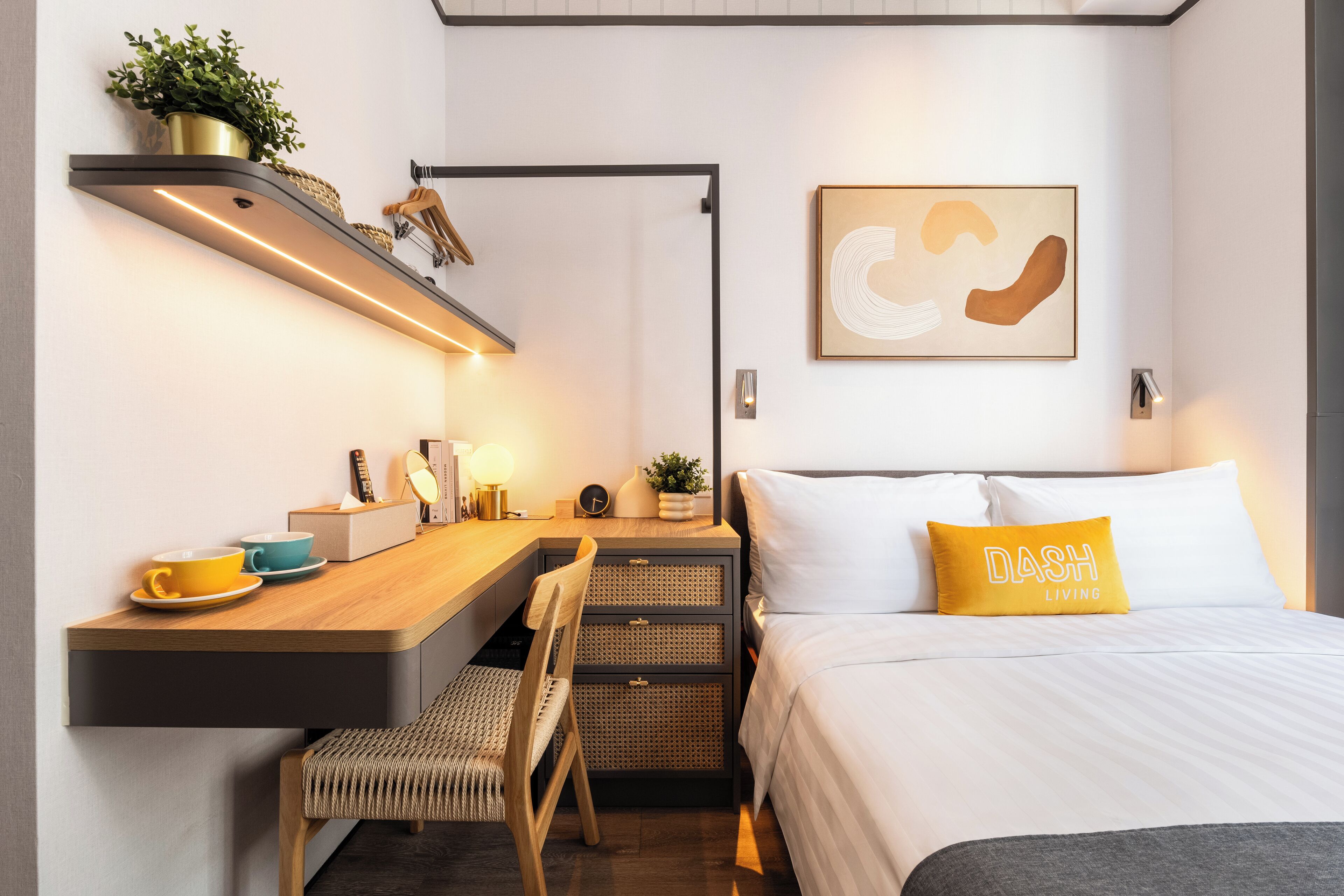 A modern hotel room with a wooden desk, a comfortable chair, a bed with a white and yellow pillow, and a wall-mounted shelf with a plant.