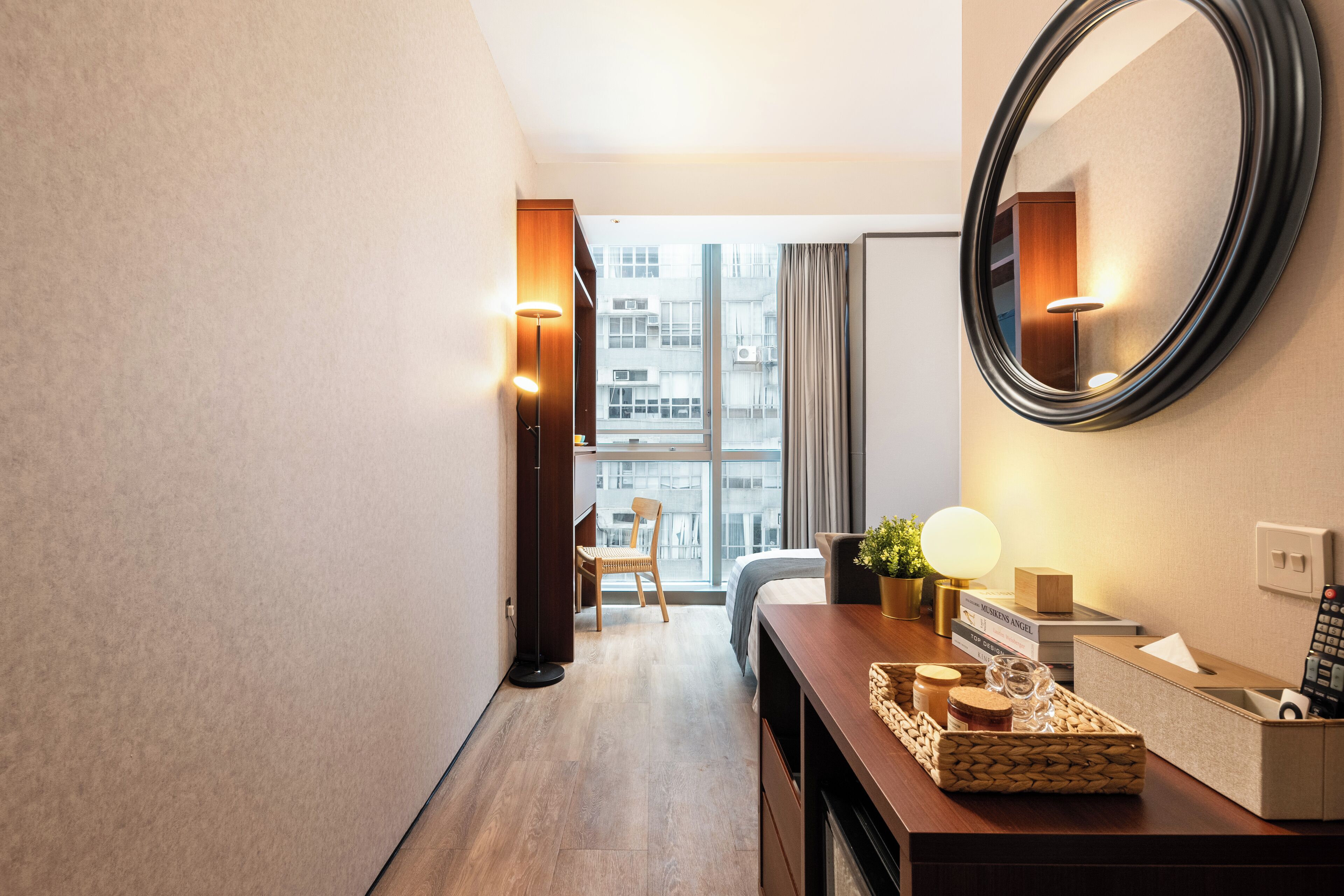 A hotel room with a wooden floor, a large mirror, a bedside table with a lamp, a chair, and a view of the city through a large window.