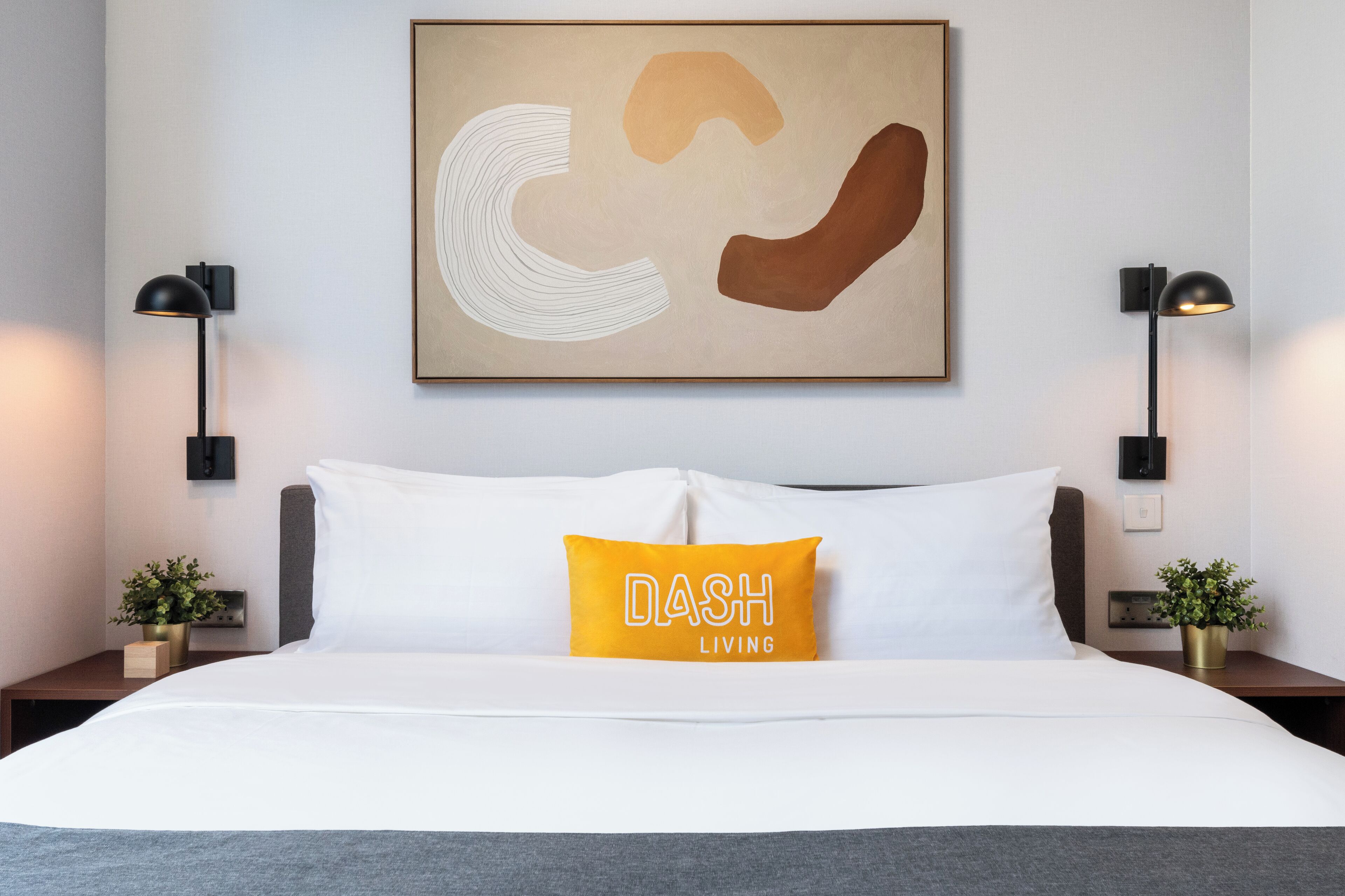 Camera d'albergo con un letto, un quadro astratto incorniciato, due lampade sui comodini e una pianta in vaso.