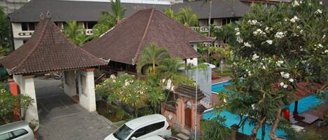 A residential area with traditional architecture, including a house with a tiled roof and a small pavilion. There is a swimming pool with a blue cover, surrounded by greenery and flowering plants.
