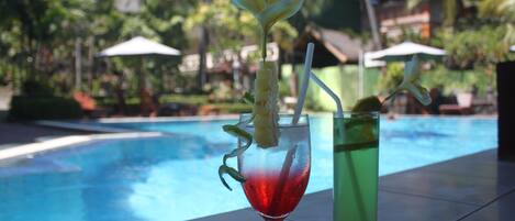 Une scène au bord de la piscine, avec deux boissons sur un plateau, une rouge et une verte, et en arrière-plan, un décor tropical comprenant des palmiers et un bâtiment.