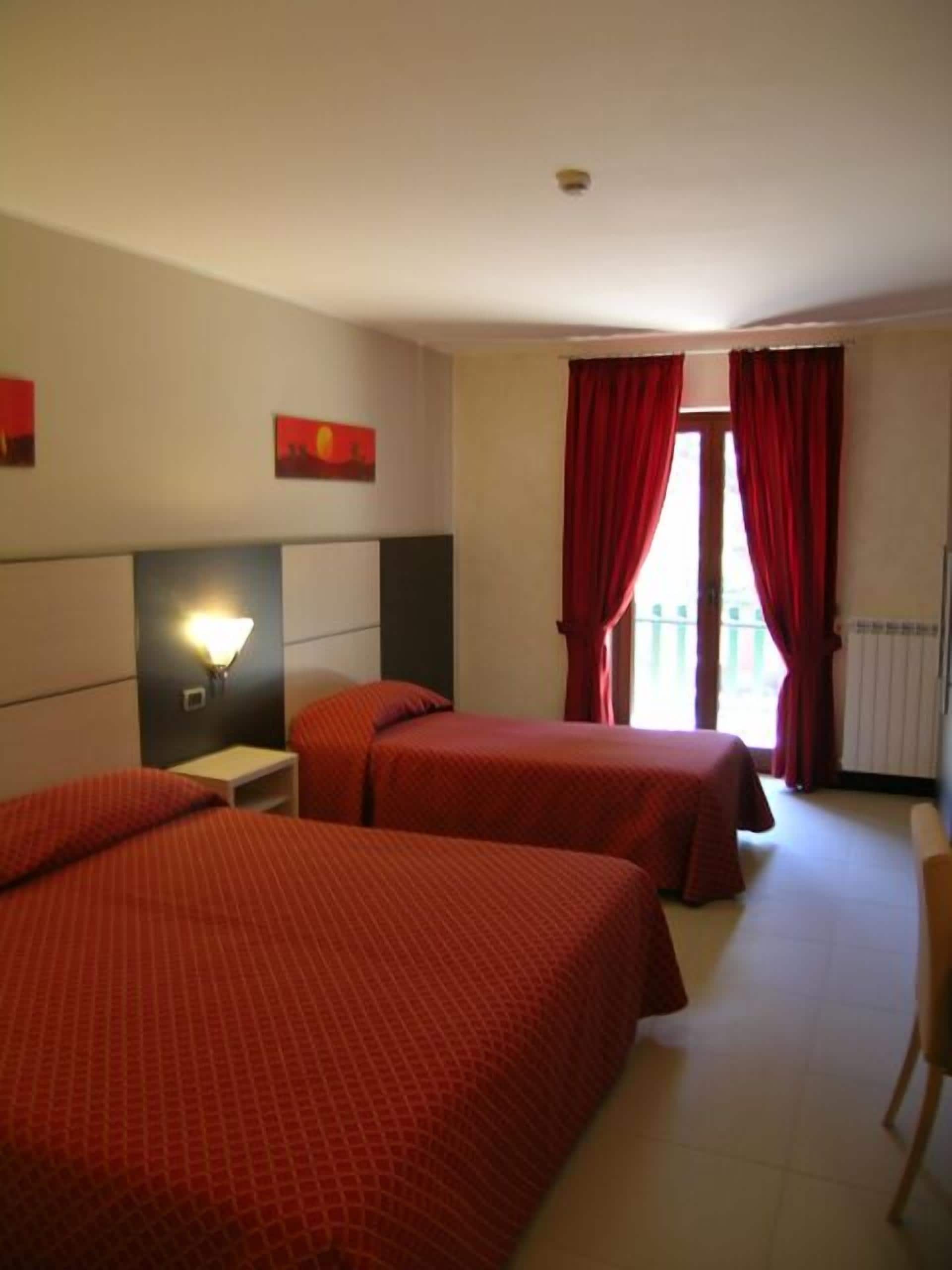 A hotel room with two beds, a nightstand, a wall-mounted lamp, and a window with red curtains.
