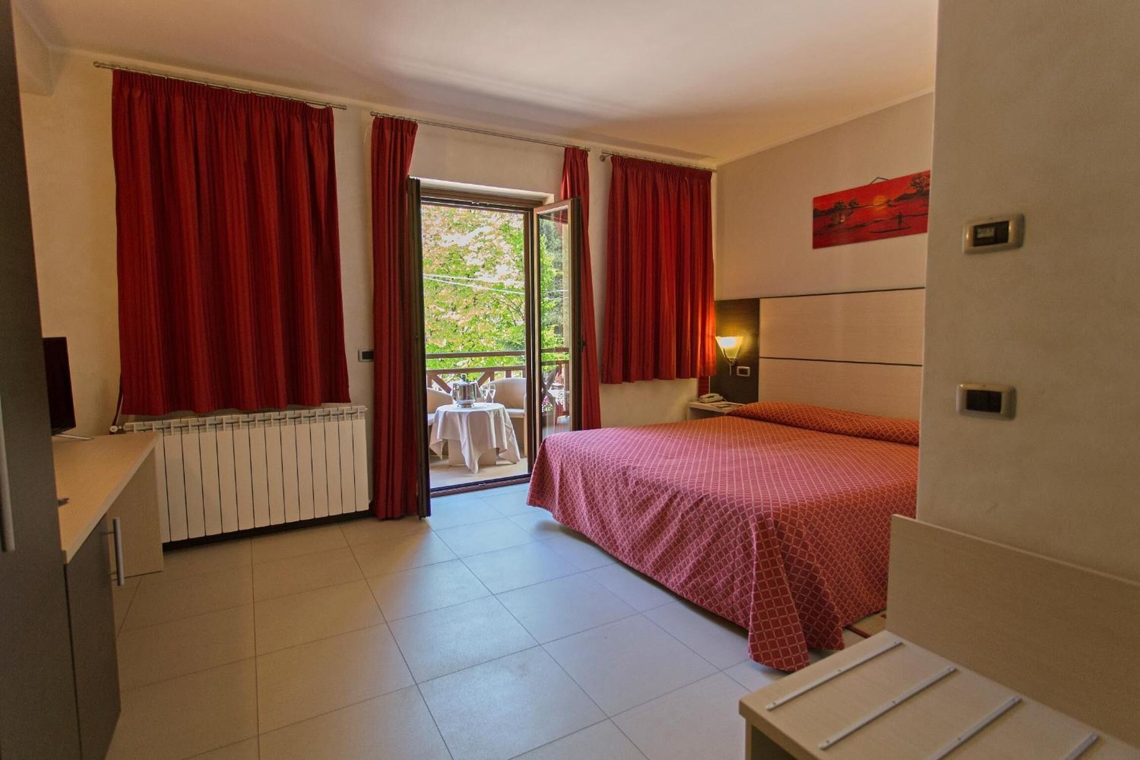 A bedroom with a bed, a nightstand, a window with red curtains, a radiator, and a view of an outdoor table and chairs.