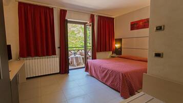 A bedroom with a bed, a nightstand, a window with red curtains, a radiator, and a view of an outdoor table and chairs.