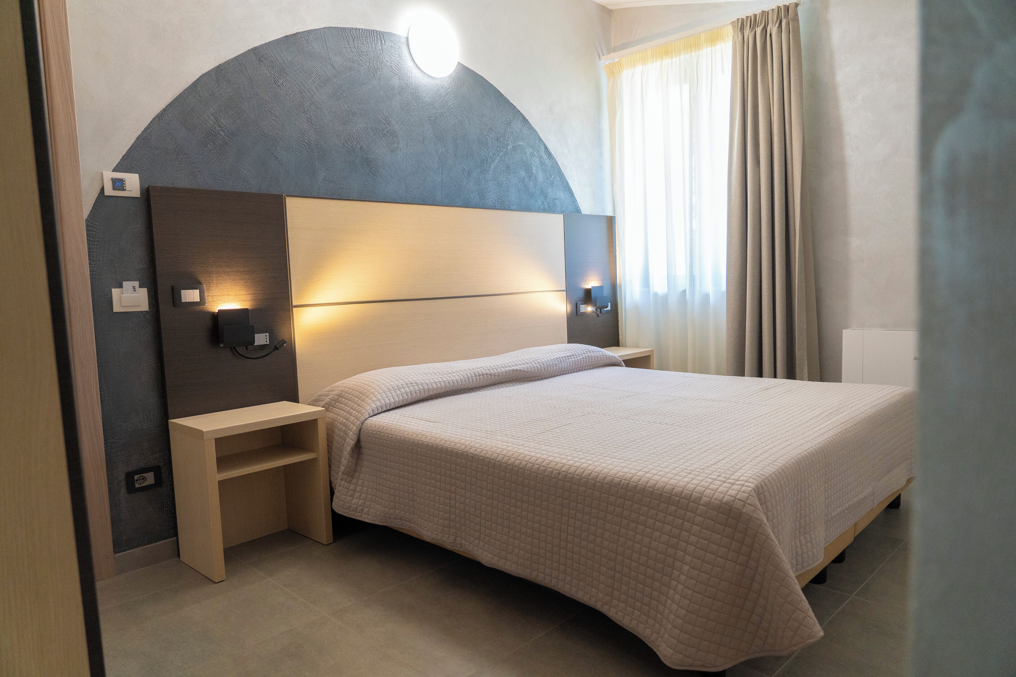 Una habitación de hotel moderna con una cama grande, un banco empotrado, un cabecero curvo y luces de lectura fijadas a la pared.