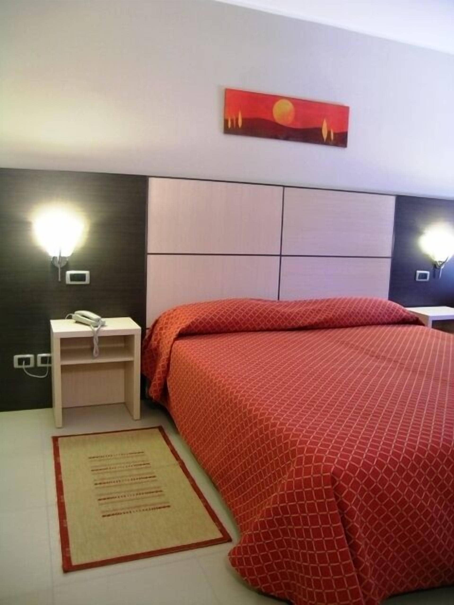 A hotel room with a bed, a nightstand with a phone, a wall-mounted light, and a painting above the bed.