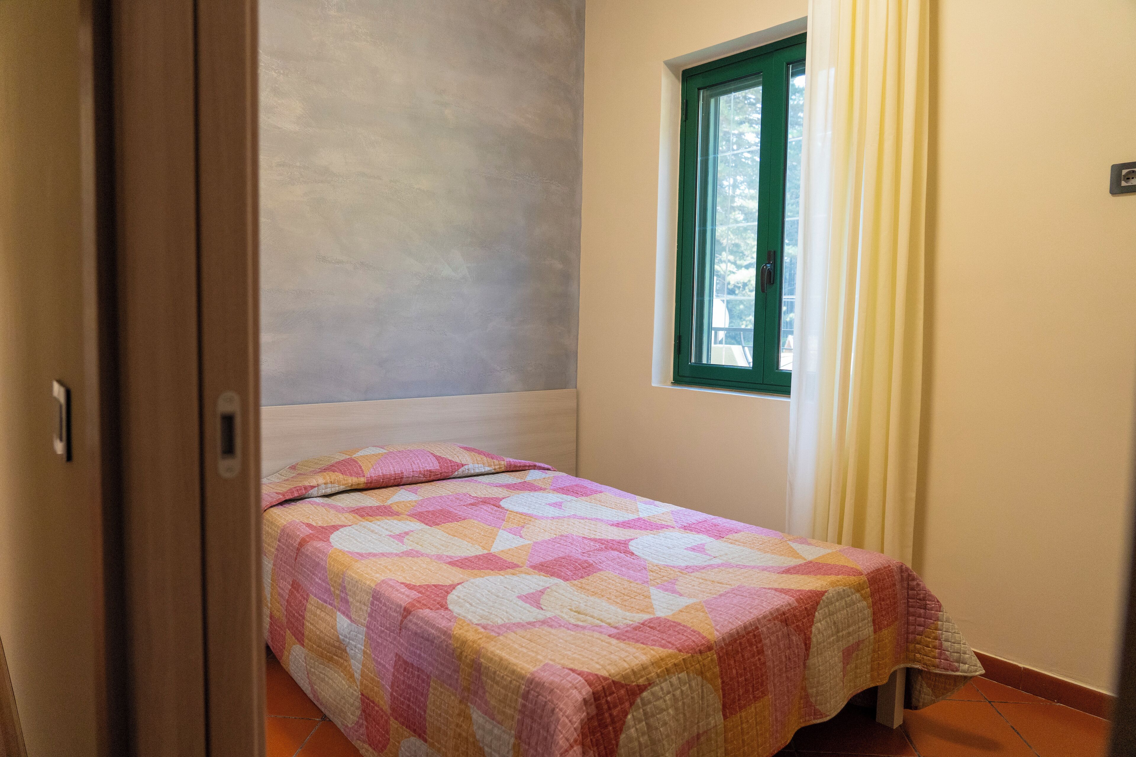 Ein kleines Zimmer mit einem Einzelbett, einer farbenfrohen Bettdecke, einem Fenster mit Vorhängen und einer schlichten Wand.