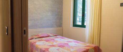 A small, single-bed room with a colorful quilt, a window with curtains, and a plain wall.