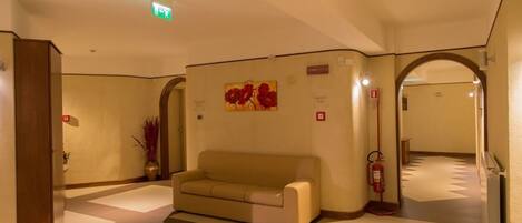 A lobby with a beige sofa, a fire extinguisher, a potted plant, and a painting on the wall.