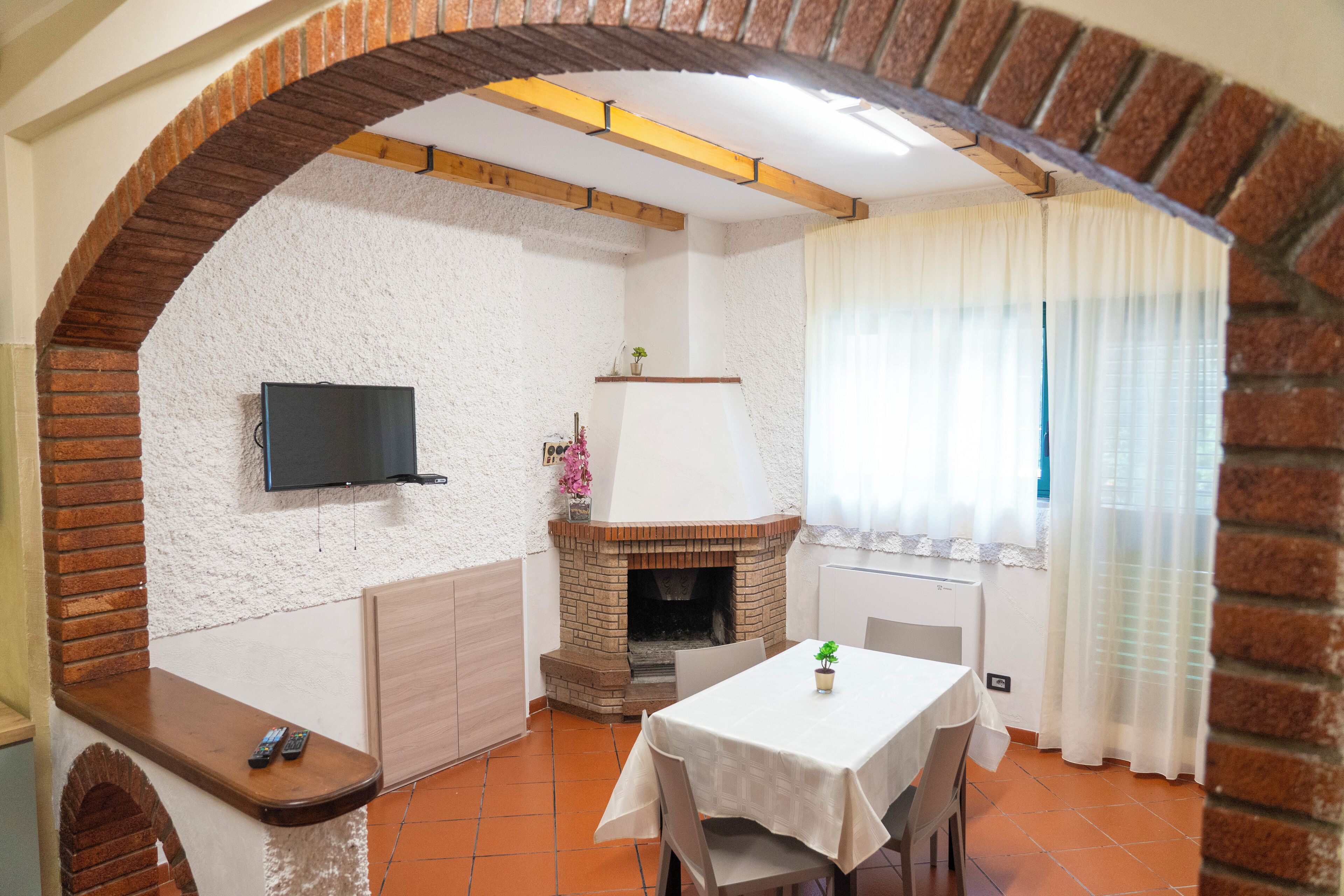 A room with a brick fireplace, a TV mounted on the wall, a table with chairs, and a window with curtains.