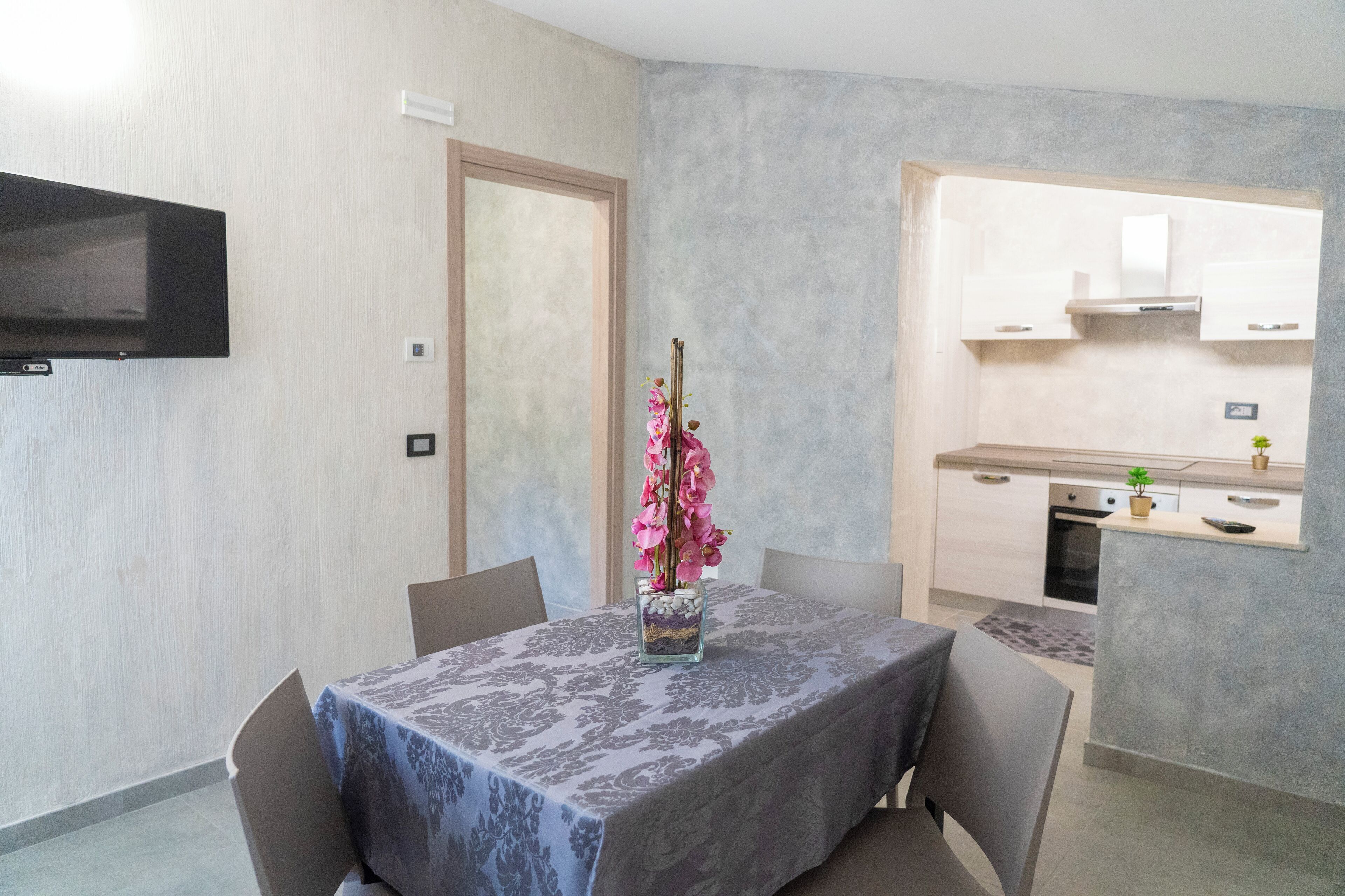 A dining area with a table set for two, a vase with pink flowers, and a kitchen in the background.