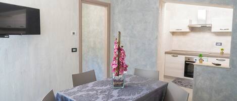 A dining area with a table set for two, a vase with pink flowers, and a kitchen in the background.