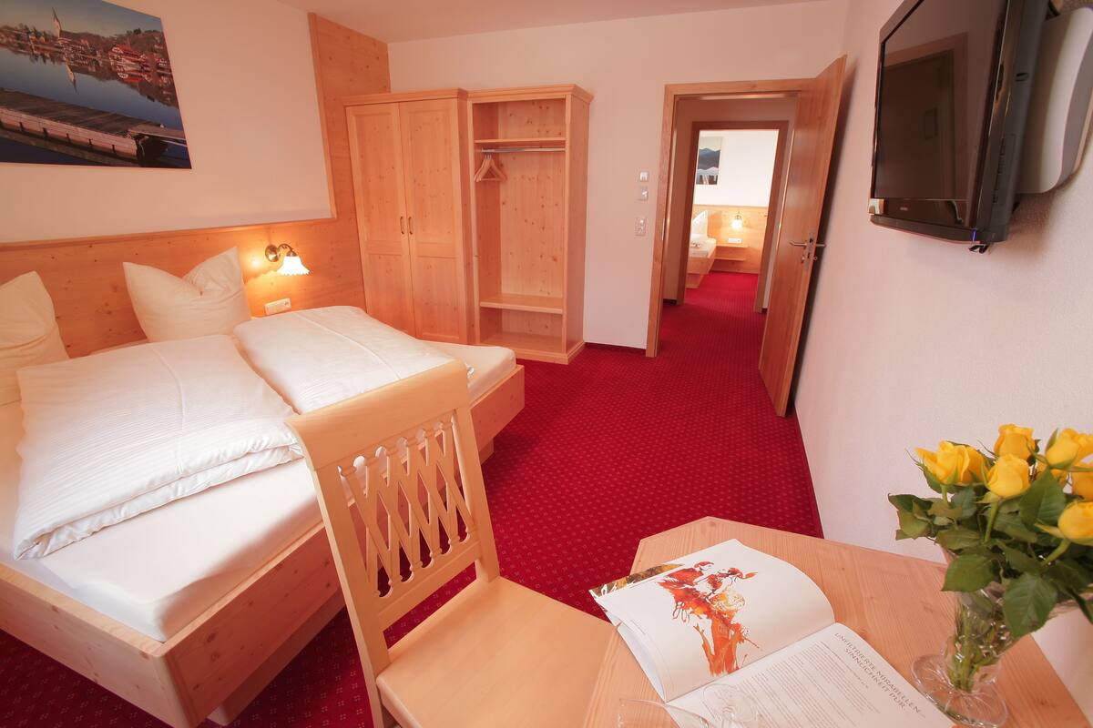 A hotel room with a bed, a wooden wardrobe, a desk with a book and a vase of flowers, and a television mounted on the wall.