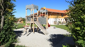 A playground with a wooden climbing structure and a slide, surrounded by a gravel area and greenery. - Pension Schweizerhaus (Weyarn)