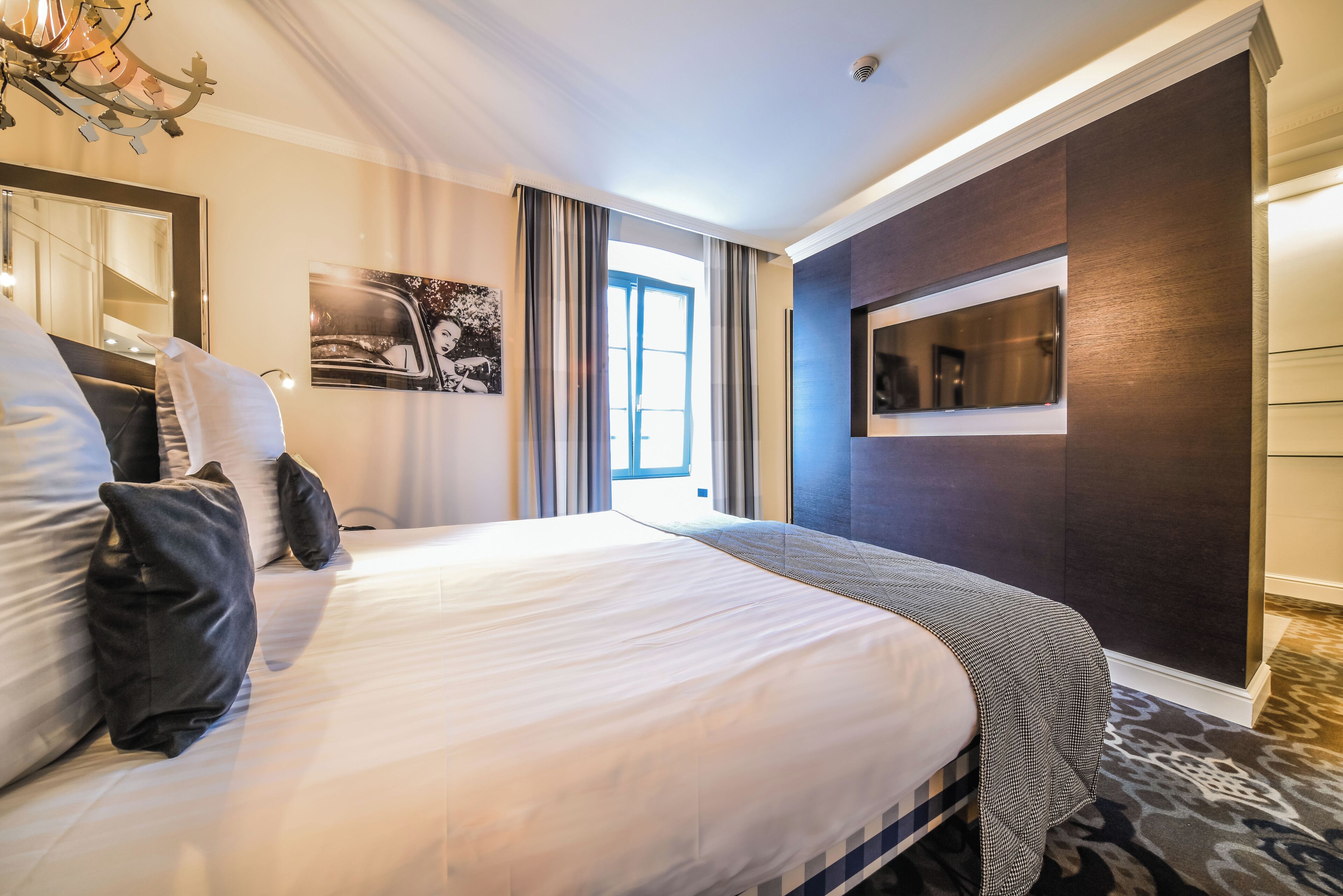 Habitación de hotel con una cama grande, un espejo, un cuadro, una ventana con cortinas y un televisor empotrado.