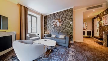 Moderni hotellihuone, jossa on harmaa sohva, valkoinen nojatuoli, pieni pyöreä pöytä ja kuviollinen matto.