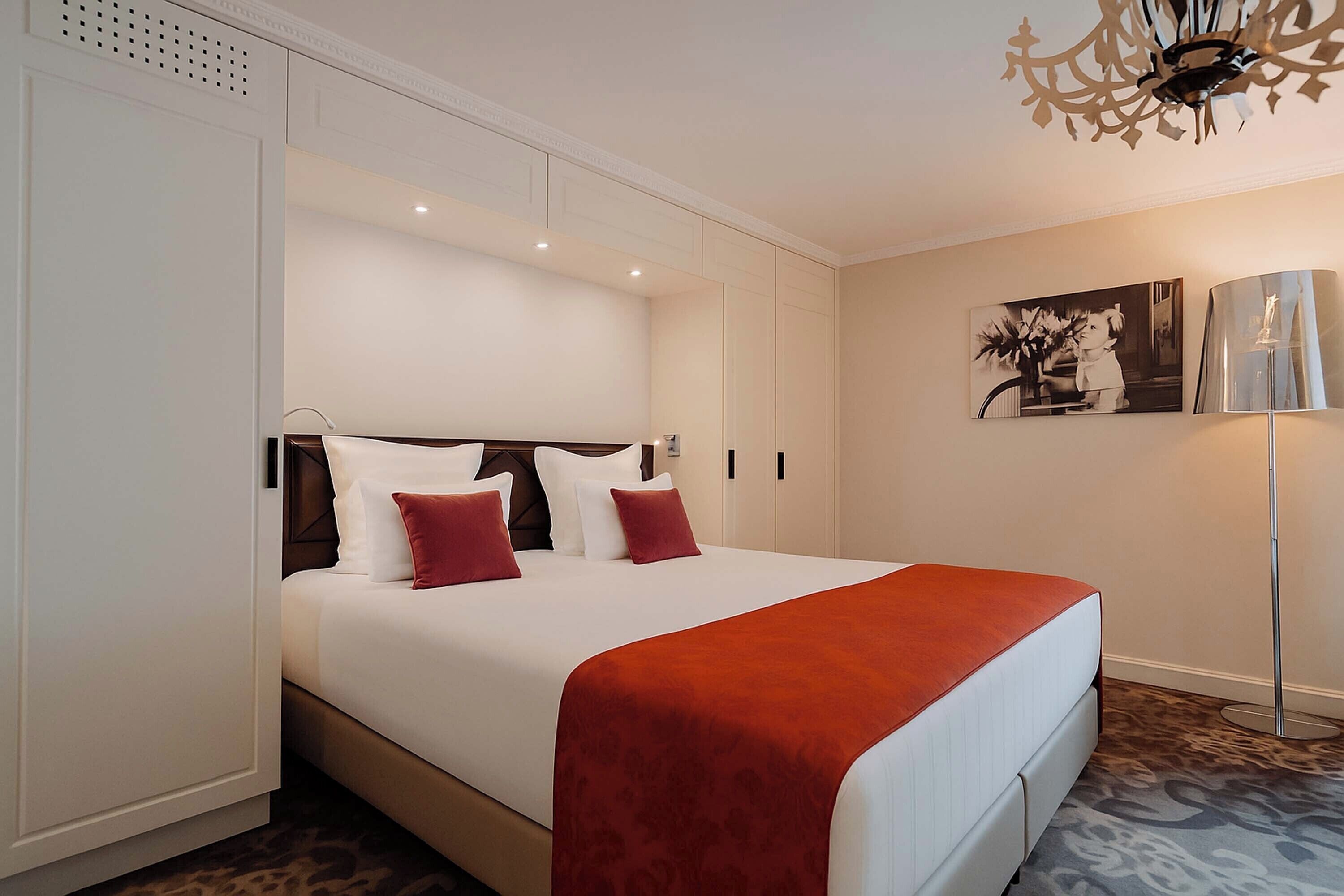 A hotel room with a bed, a red throw blanket, a floor lamp, and a painting on the wall.