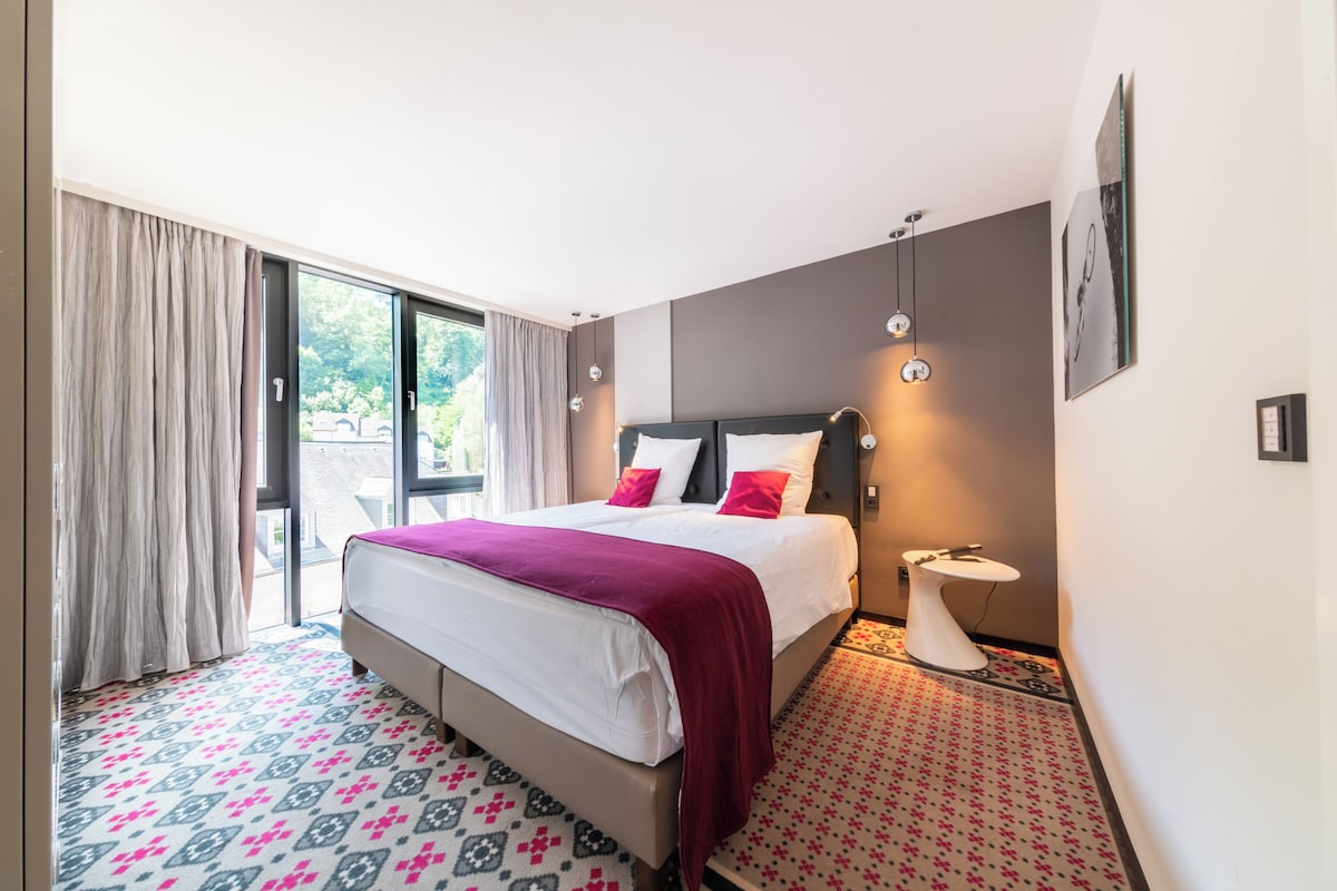 A hotel room with a bed, a bedside table, a window with curtains, and a patterned floor.