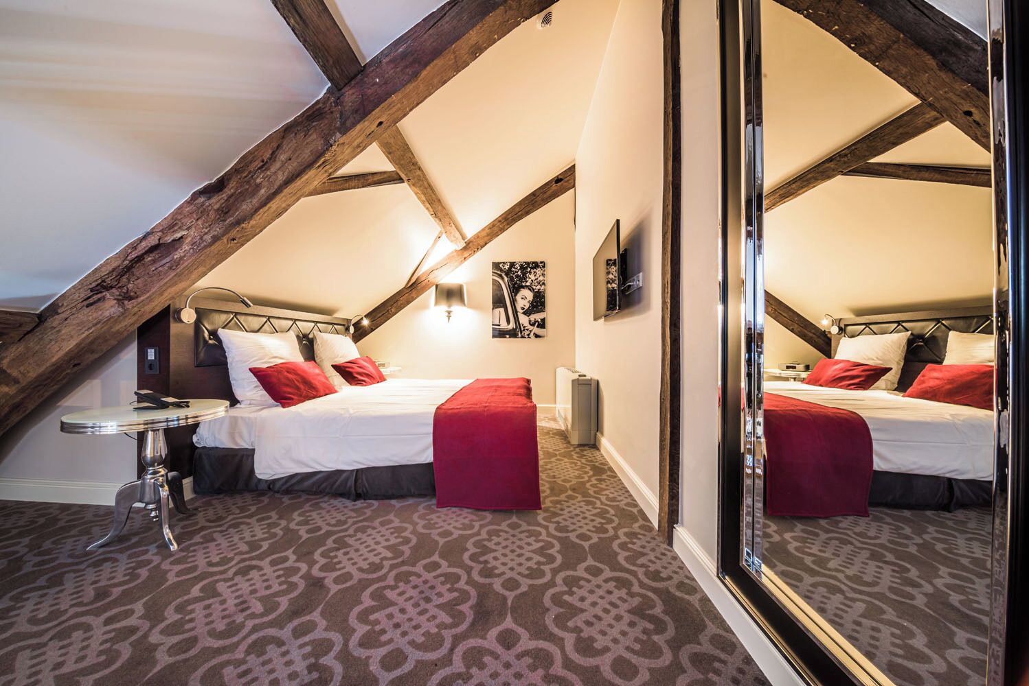 Een slaapkamer met een schuine plafond, een bed met rode en witte beddengoed, een klein rond tafeltje en een grote spiegel.