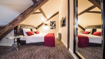Een slaapkamer met een schuine plafond, een bed met rode en witte beddengoed, een klein rond tafeltje en een grote spiegel.
