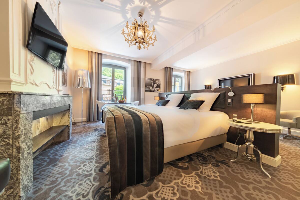 A hotel room with a large bed, a flat-screen TV mounted on the wall, a chandelier, and a patterned carpet.