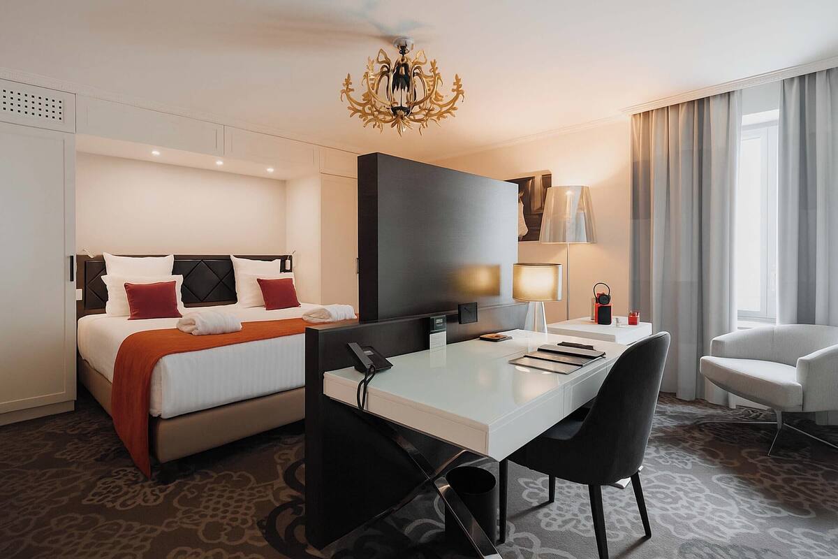 A hotel room with a bed, a desk, a chair, a TV, and a chandelier.