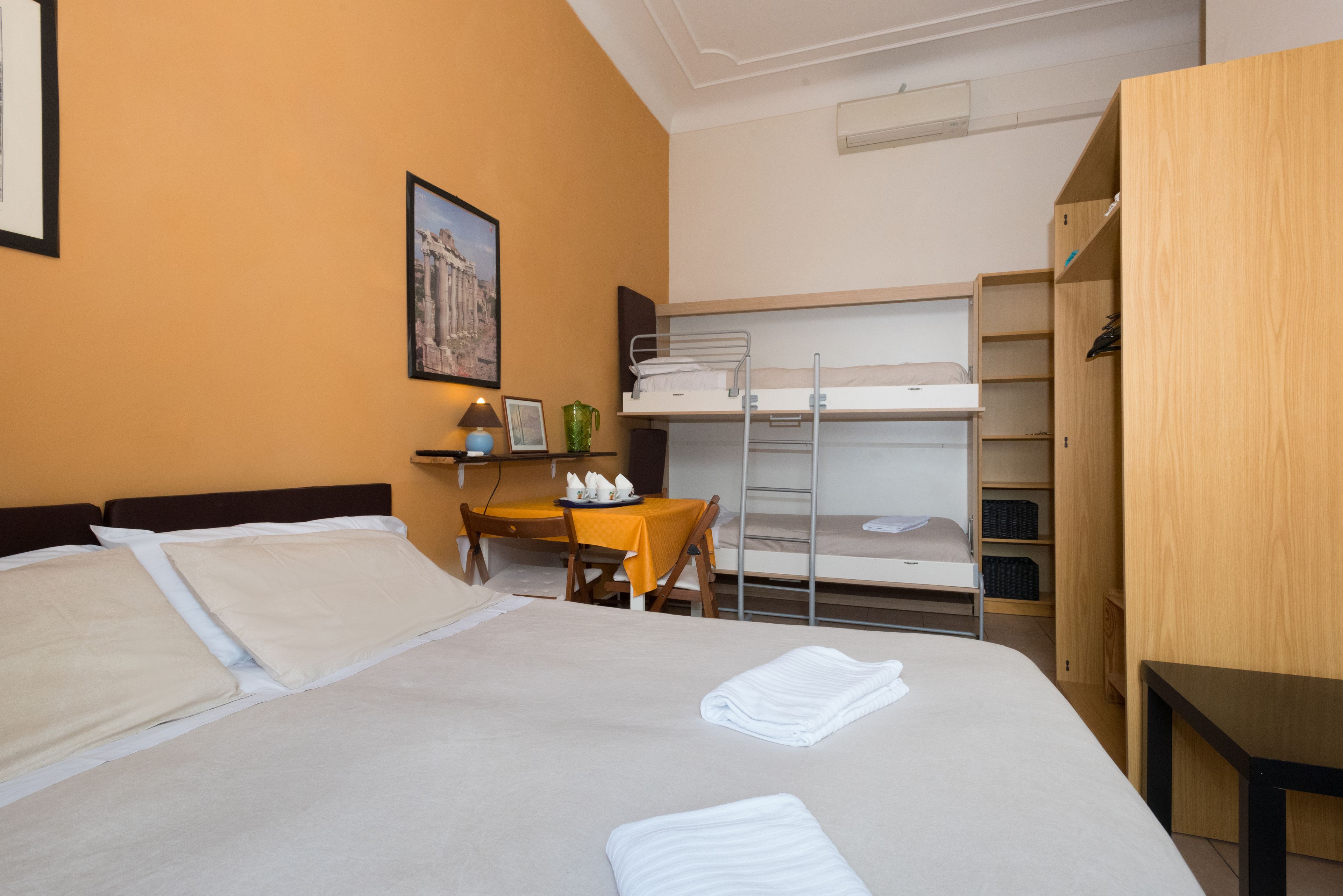 A hotel room with a bed, a desk, a chair, a small table, a bunk bed, and a wardrobe.