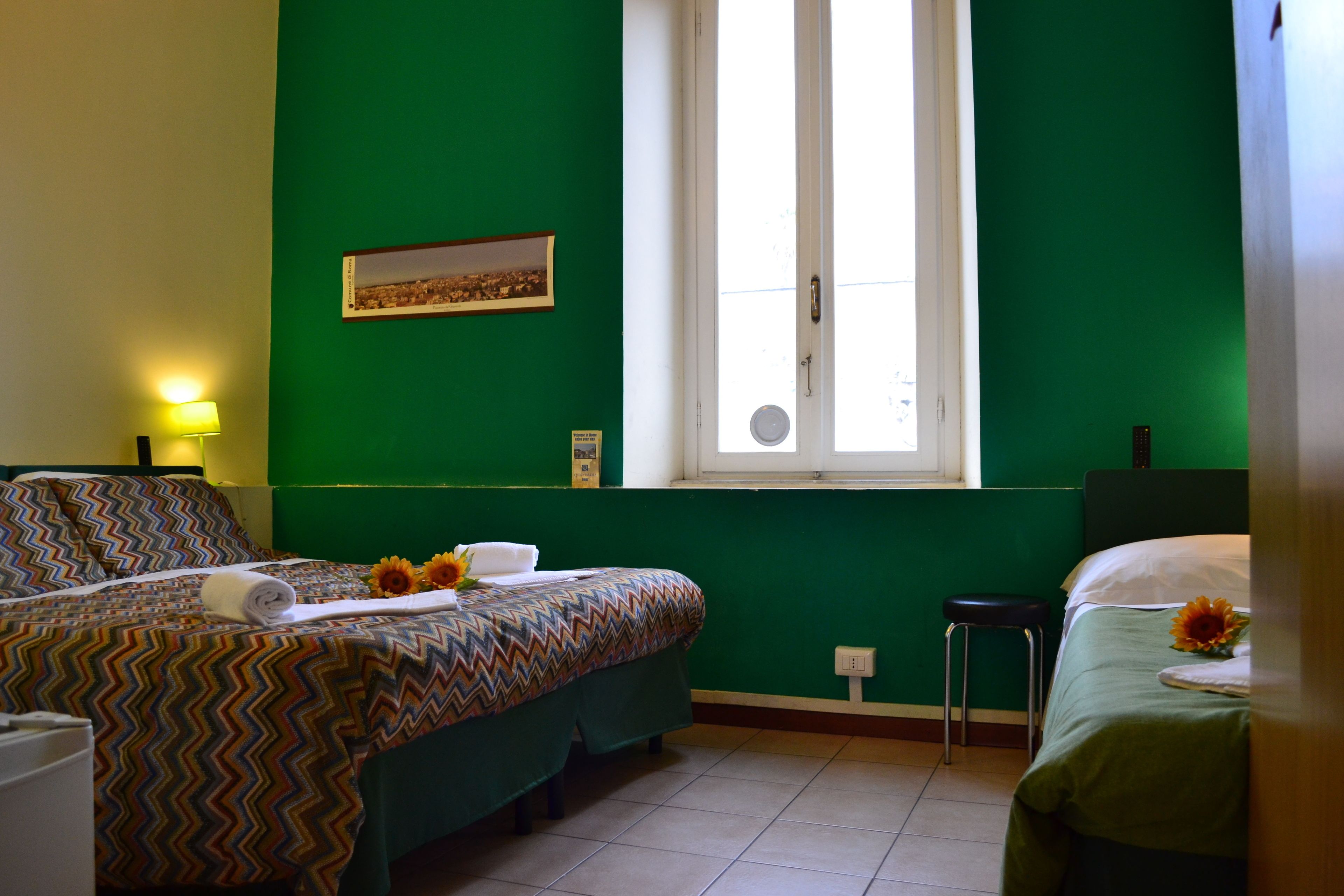 Habitació d'hotel amb dos llits, una finestra, un quadre a la paret i una tauleta de nit amb una làmpada.