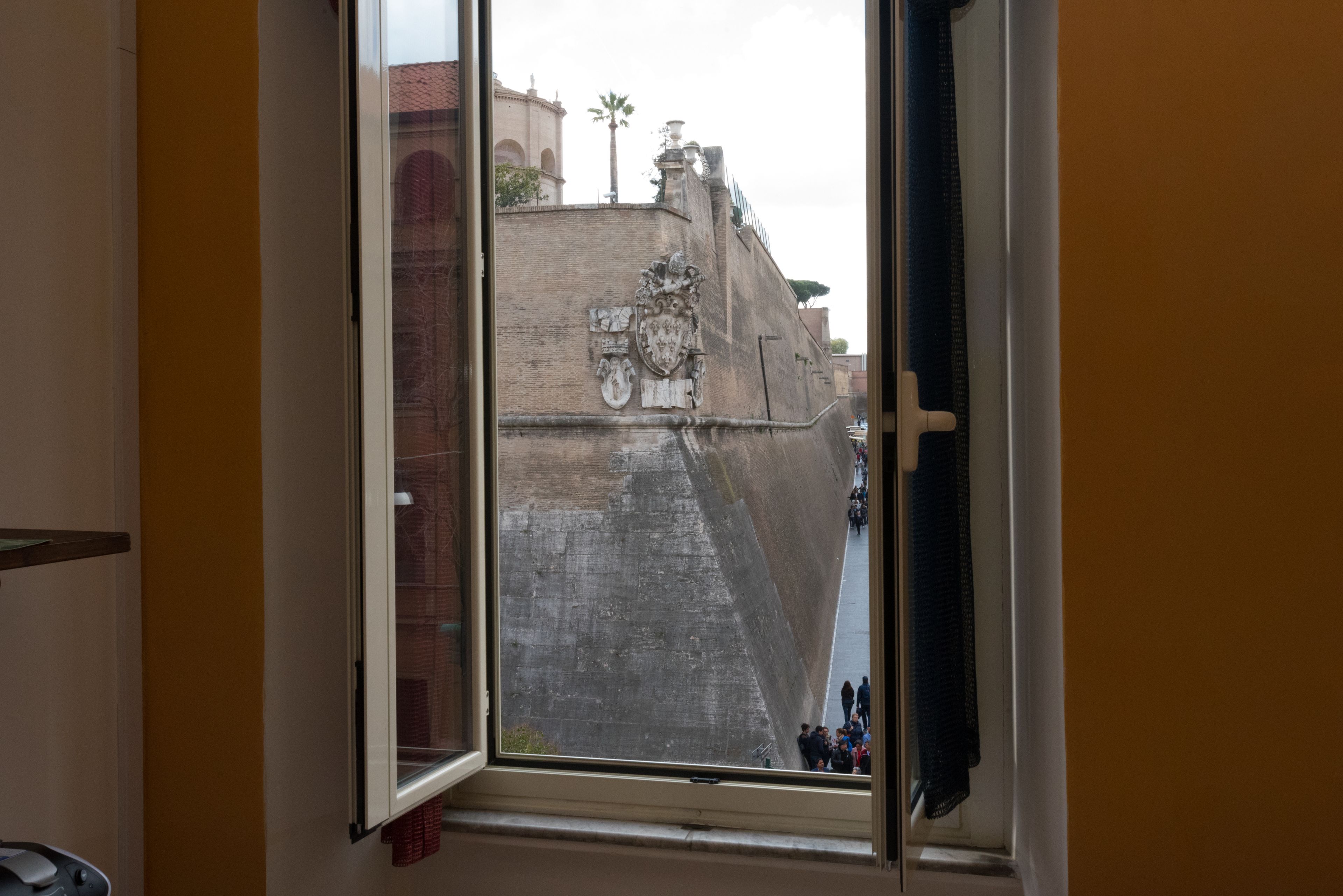 Une vue par la fenêtre sur un mur de pierre historique orné de blasons, avec un palmier et un bâtiment en arrière-plan.