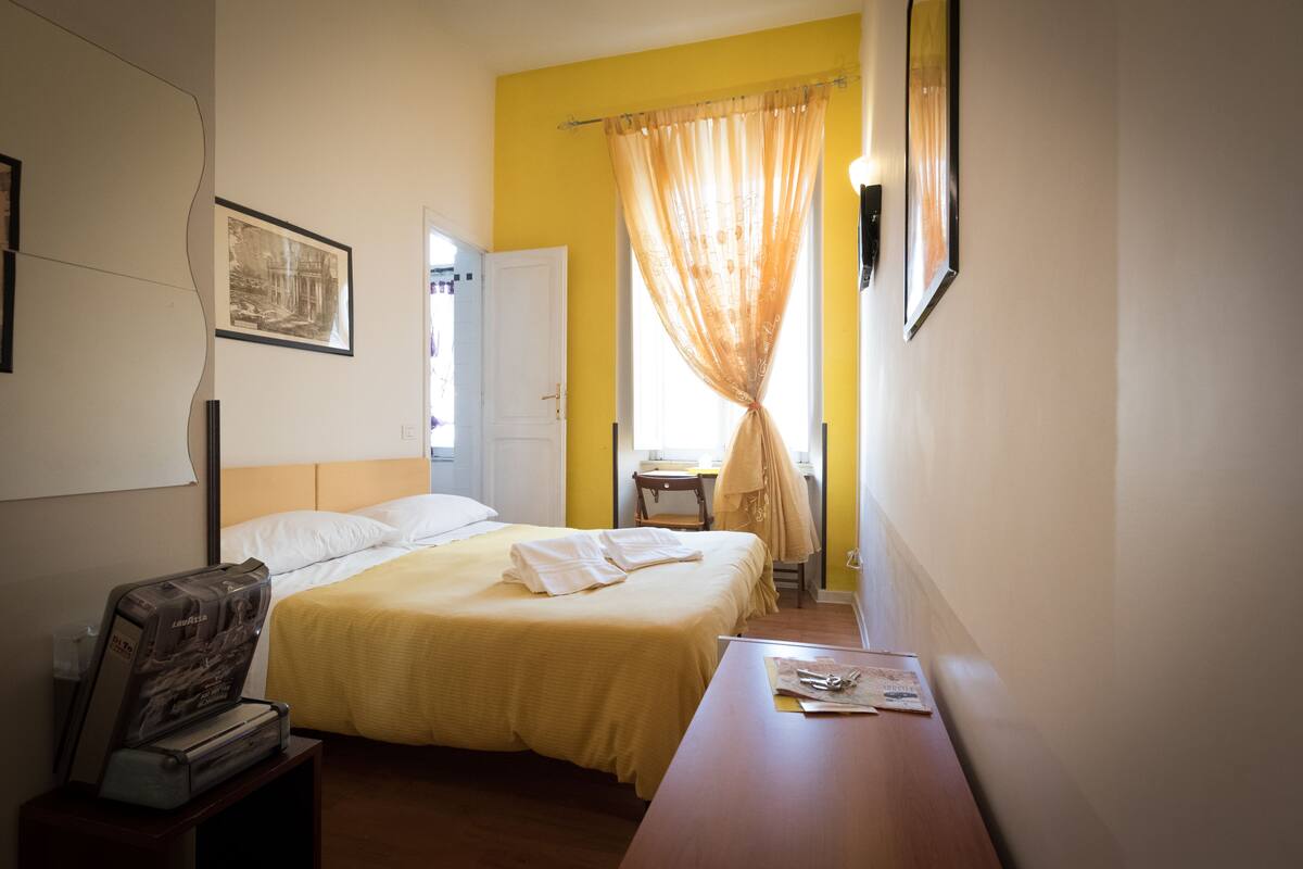 A hotel room with a bed, a desk, a chair, a window with yellow curtains, and a framed picture on the wall.