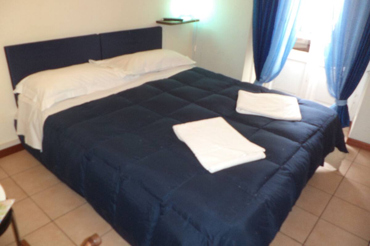 A neatly made bed with a dark blue bedspread and white linens, a bedside table with a lamp, and a window with blue curtains.
