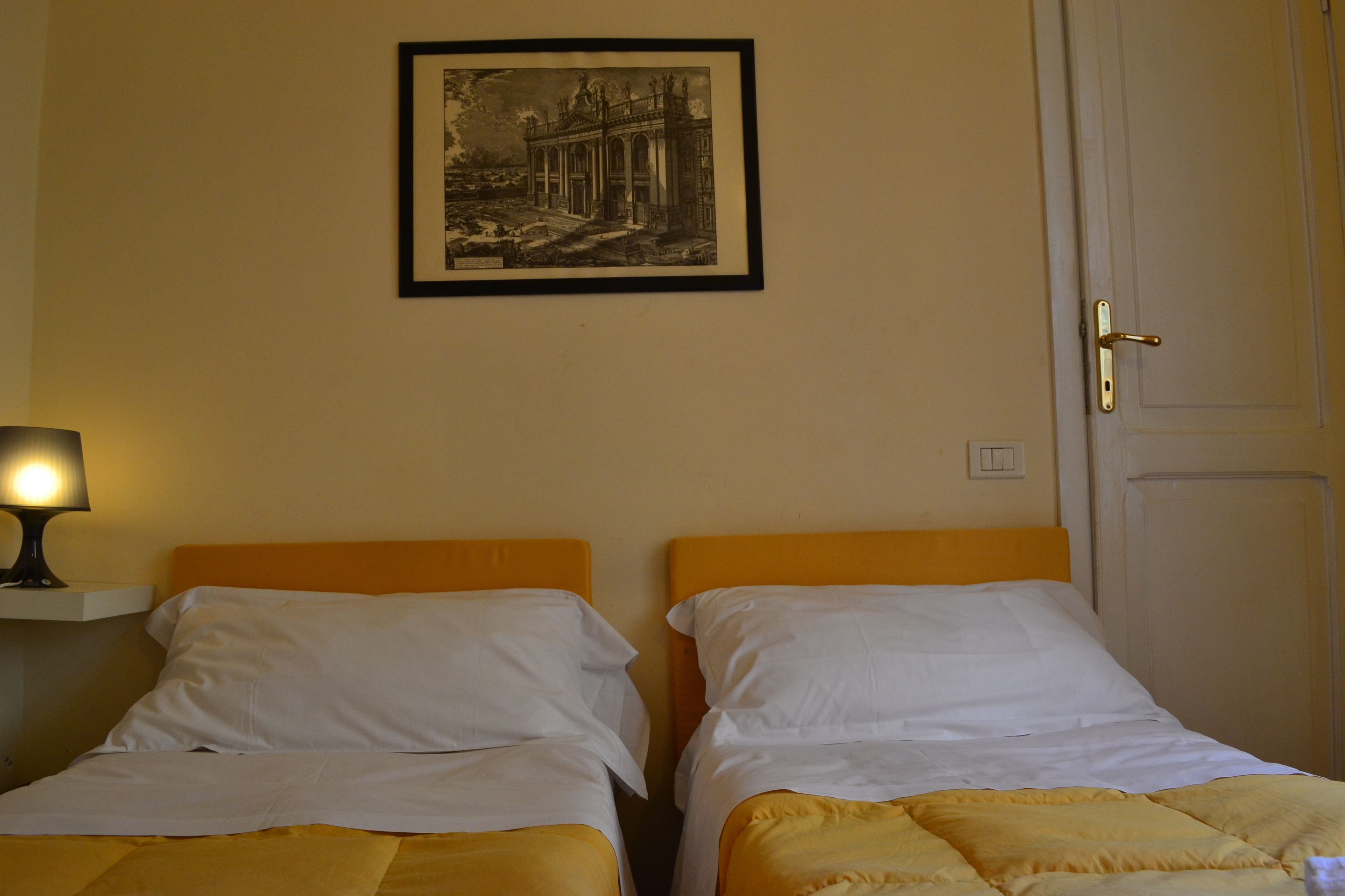 Una stanza con due letti, un quadro appeso al muro, una lampada sul comodino e una porta.
