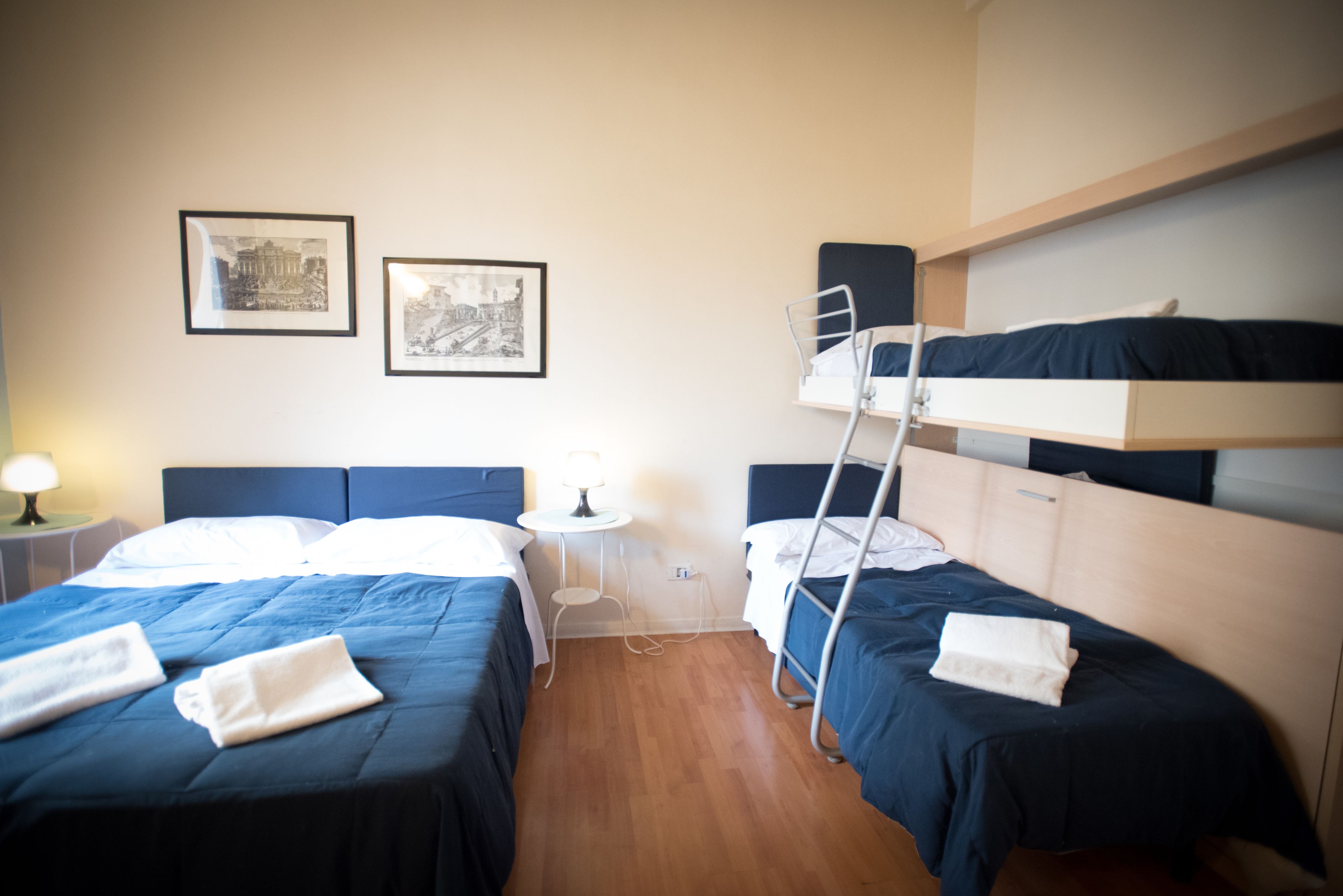Una habitación de hotel con dos camas individuales, una litera, una mesita con lámpara y dos cuadros en la pared.