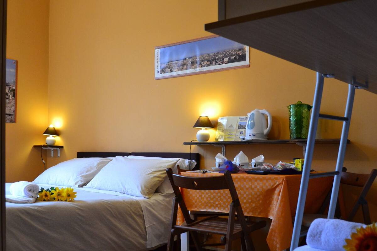 A dormitory room with bunk beds, a desk with a lamp, a chair, and a small table with a kettle and a cup.