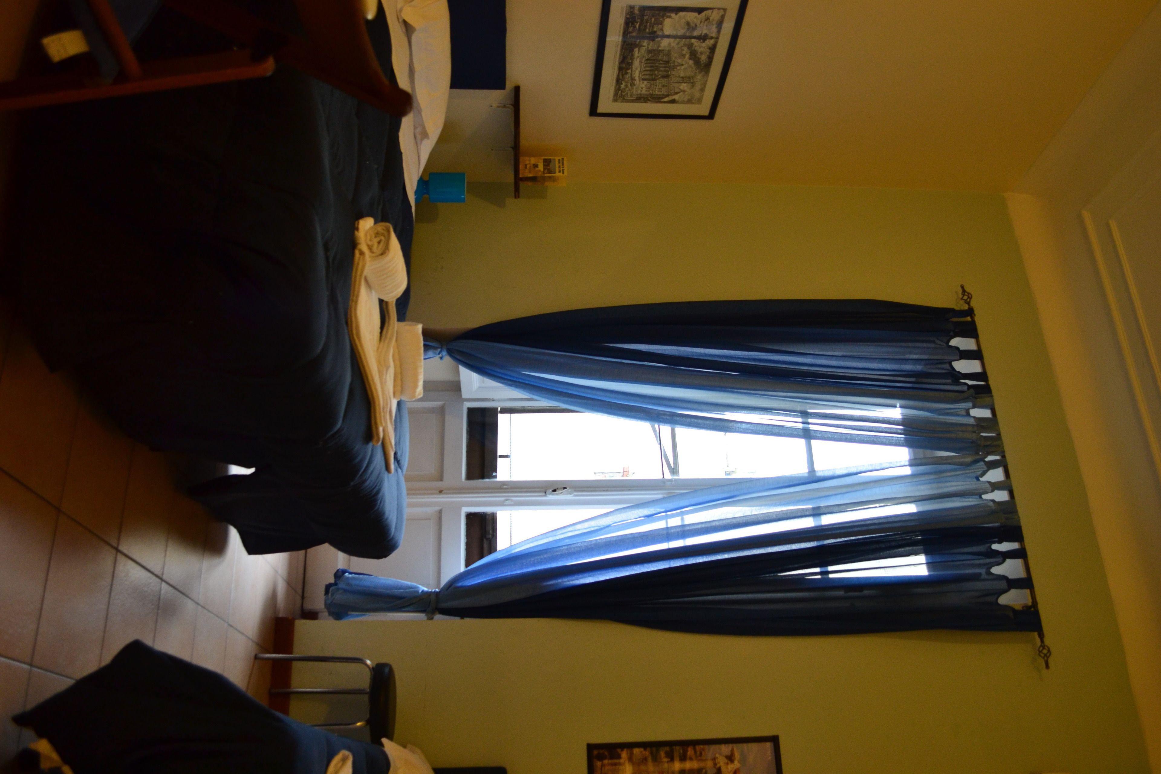 A room with a bed, a chair, a window with curtains, a framed picture on the wall, and a small table.