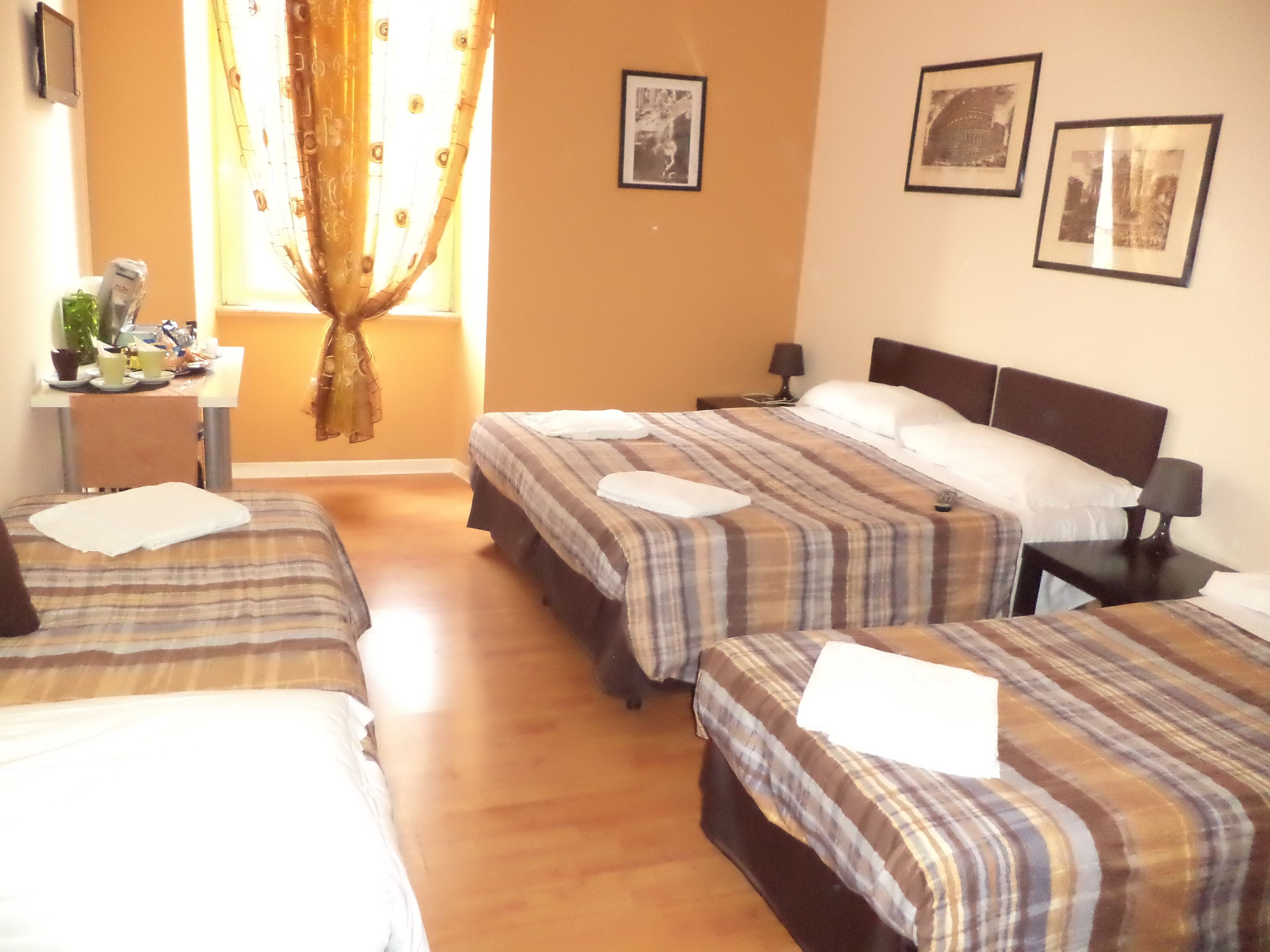 A hotel room with three beds, a desk with a chair, a window with curtains, and framed pictures on the wall.