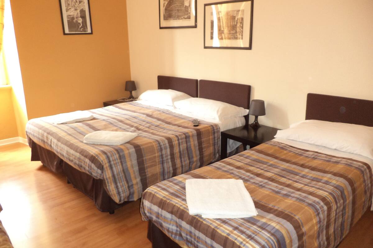 A hotel room with two beds, each with a plaid bedspread and white pillows. There are framed pictures on the wall and a small table with a lamp between the beds.