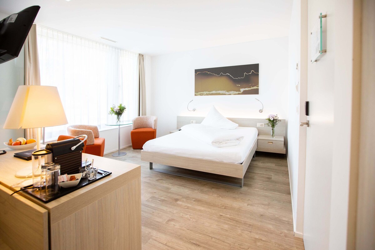 A modern hotel room with a bed, bedside table, lamp, chair, and a desk with a laptop and coffee cup.