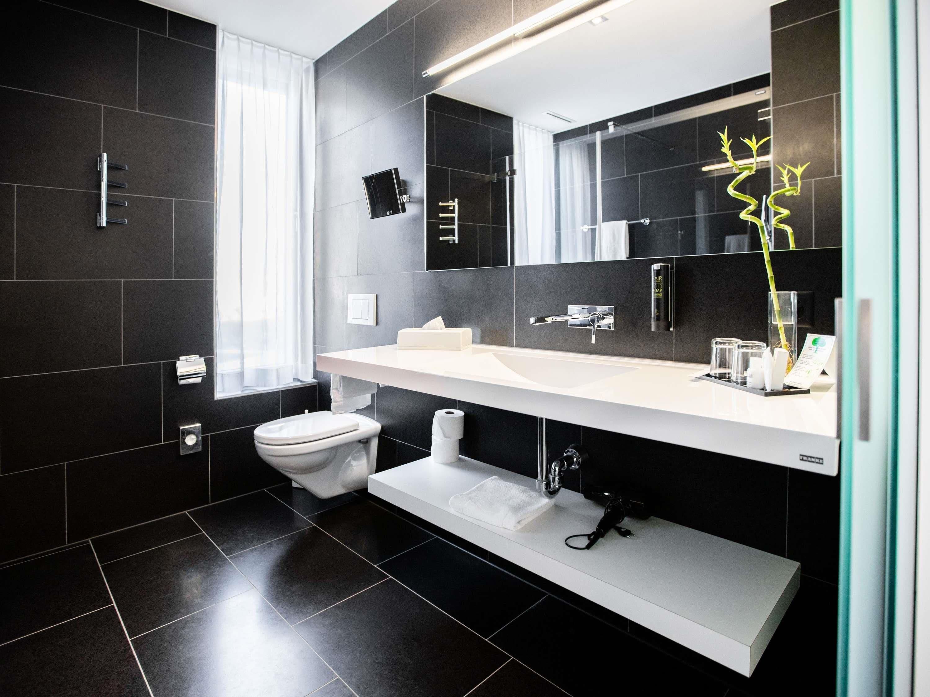 Un baño moderno con un lavamanos blanco, baldosas oscuras, un inodoro y un espejo con una planta decorativa.