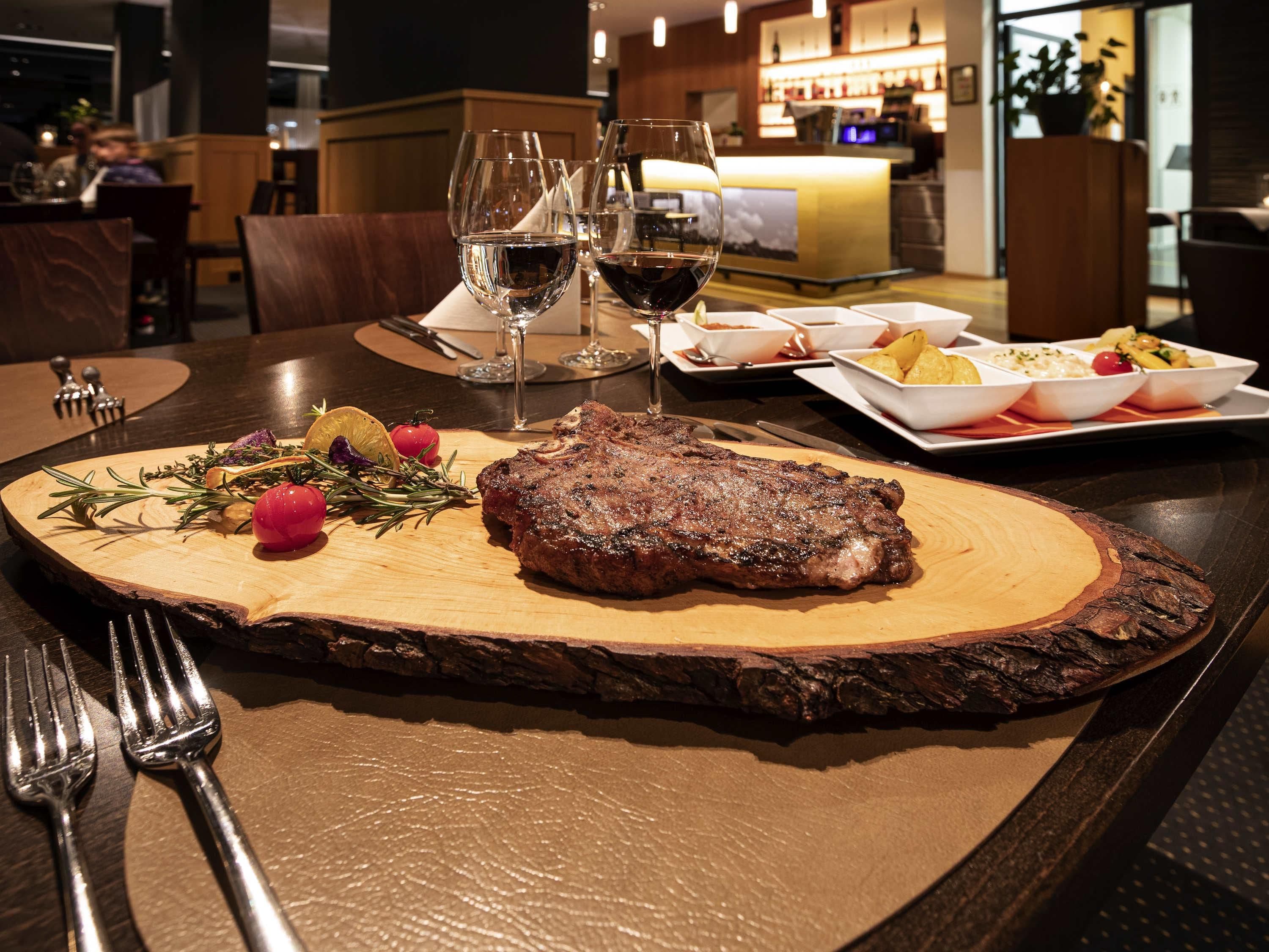 Un steak garni, présenté sur une planche en bois, des verres à vin et des assiettes remplies de nourriture sur une table dans un restaurant.
