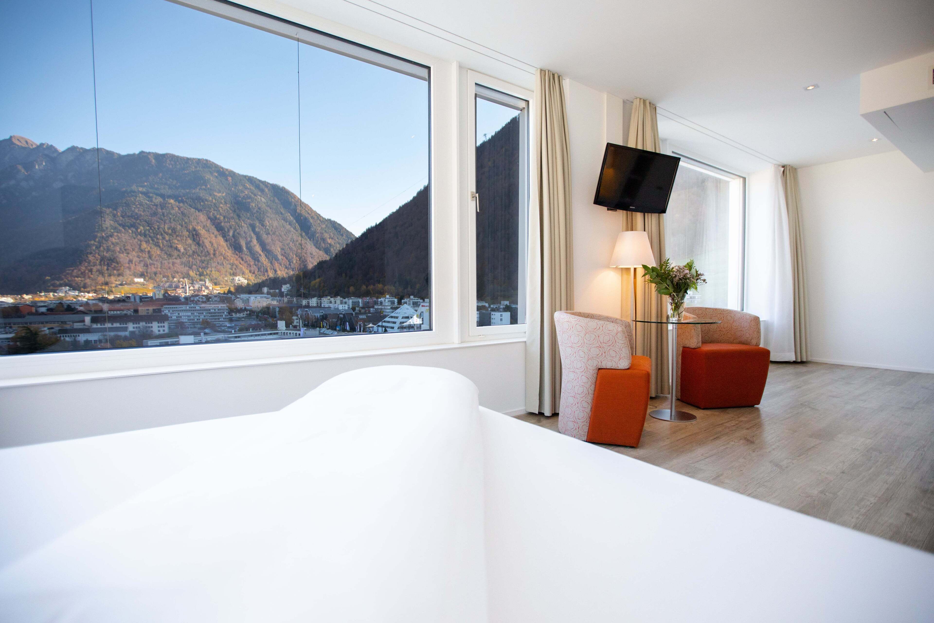 Une chambre d’hôtel moderne dotée d’une grande fenêtre offrant une vue sur les montagnes, d’un téléviseur à écran plat fixé au mur et d’un coin salon confortable avec une lampe et un vase de fleurs.