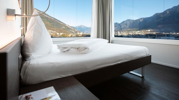A modern hotel room with a bed, large windows, and a view of mountains. - Mercure Chur City West (Chur)